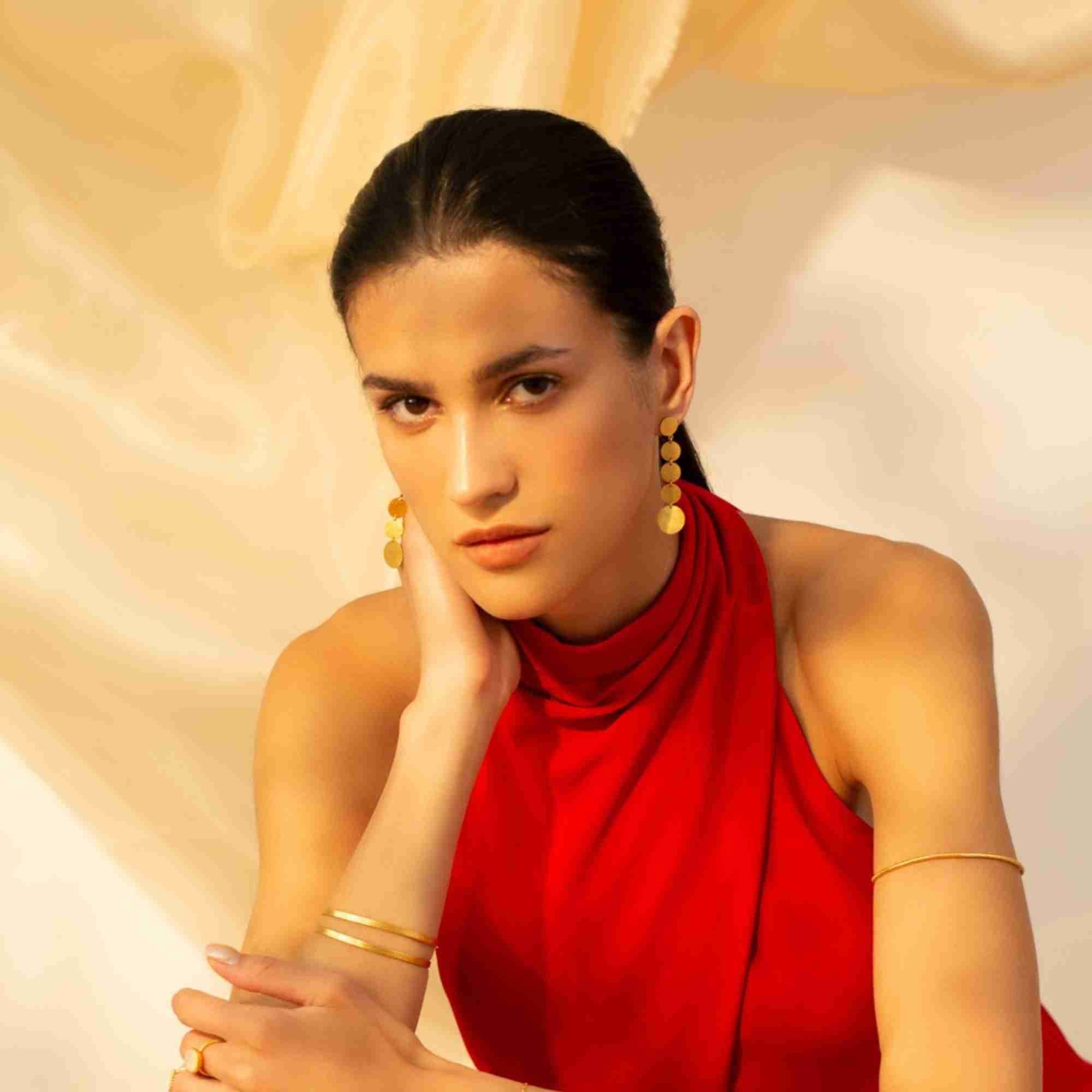 Model wearing a red halter dress styled with the gold Shimmering Disks Sculptural Flat-Band Double Cuff on her wrist, paired with matching gold drop earrings and an upper arm cuff.