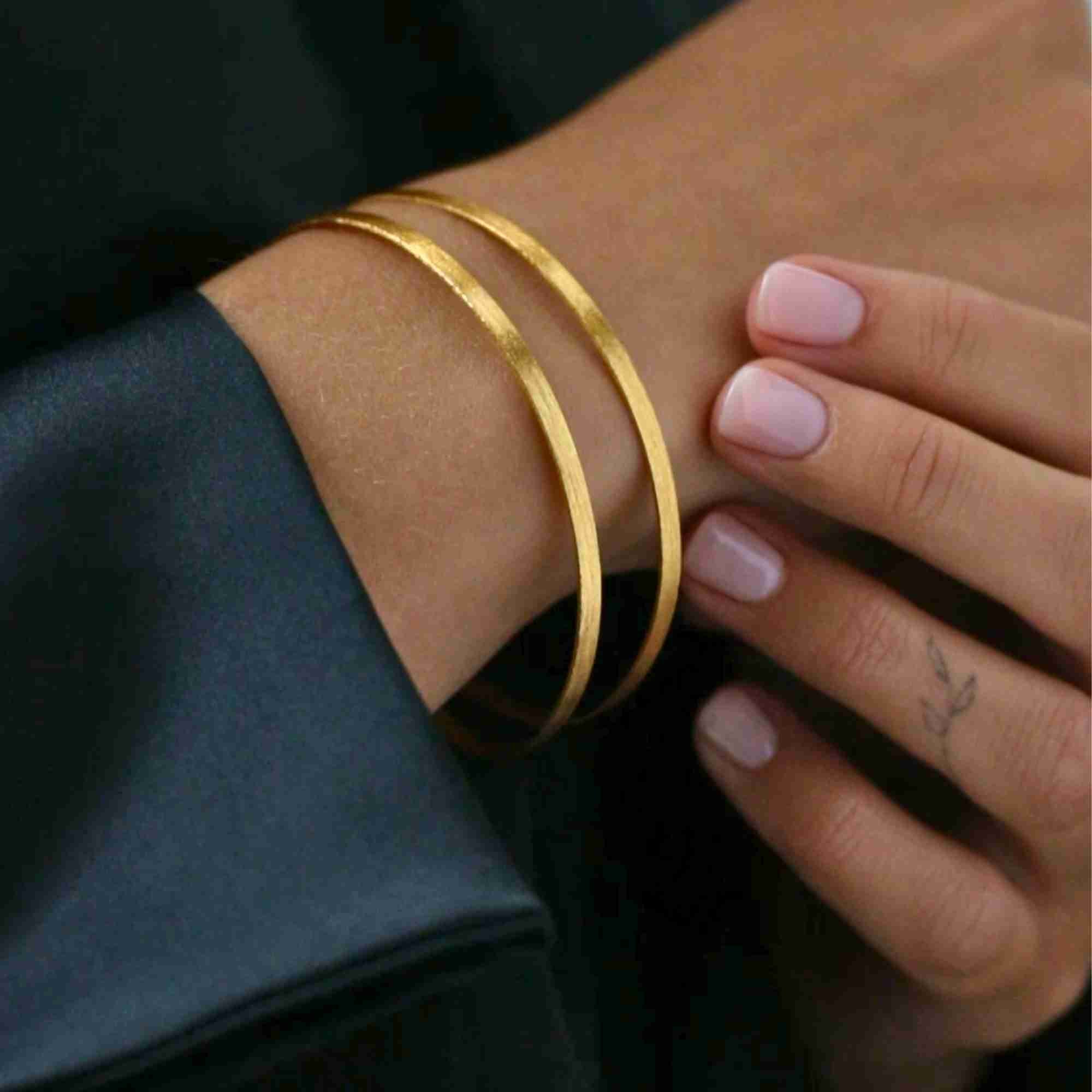 Close-up shot of a wrist adorned with the gold Shimmering Disks Sculptural Flat-Band Double Cuff, showcasing its brushed matte finish and dual parallel band structure against a black satin sleeve.