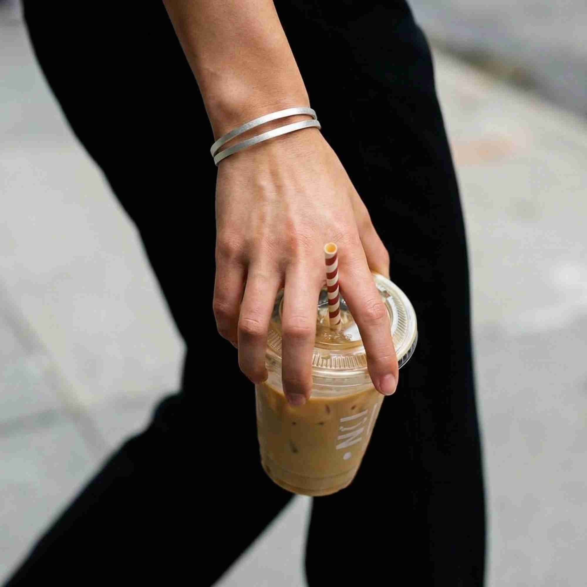 Street style shot of a model carrying an iced coffee while wearing the silver Shimmering Disks Sculptural Flat-Band Double Cuff bracelet paired with black trousers.