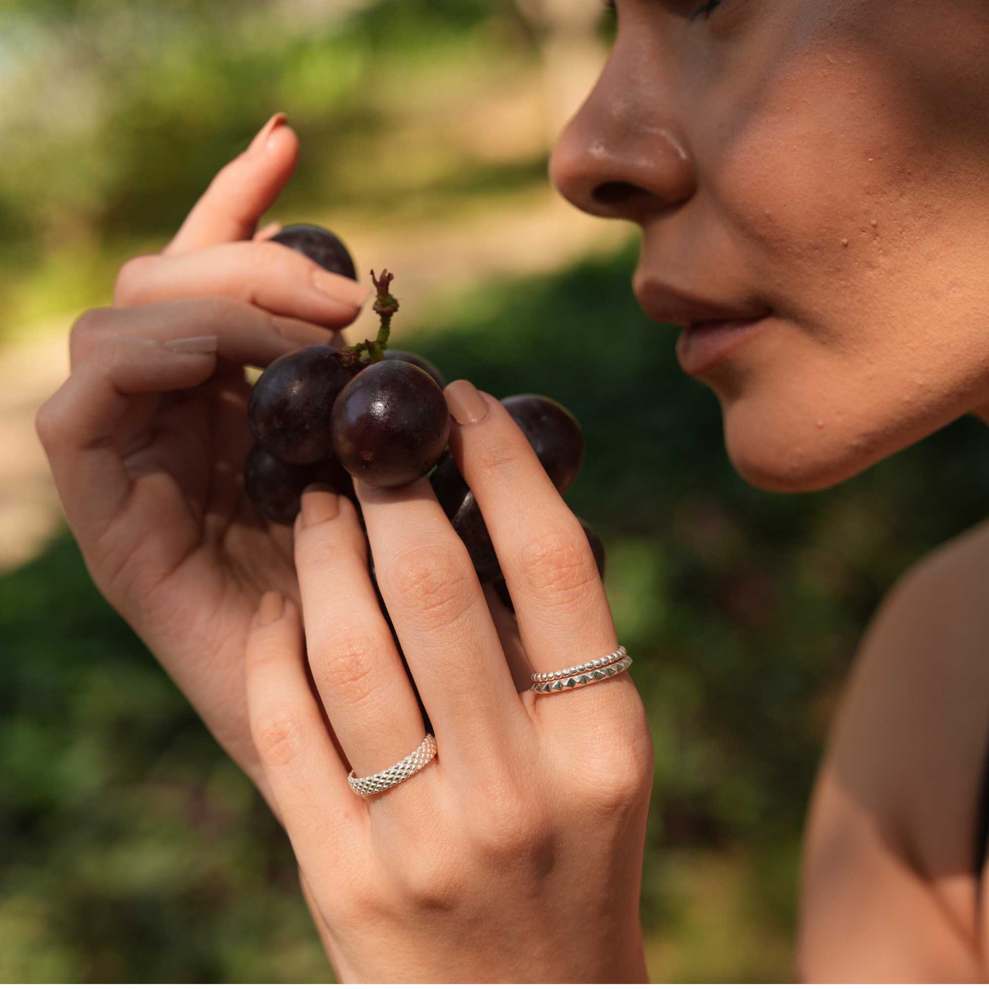Angled lifestyle shot focusing on the model's hand cradling grapes, displaying the clean lines and vintage-inspired texture of the Delicate Vintage Beaded Stacking Ring Silver, by Protsaah Zurich.