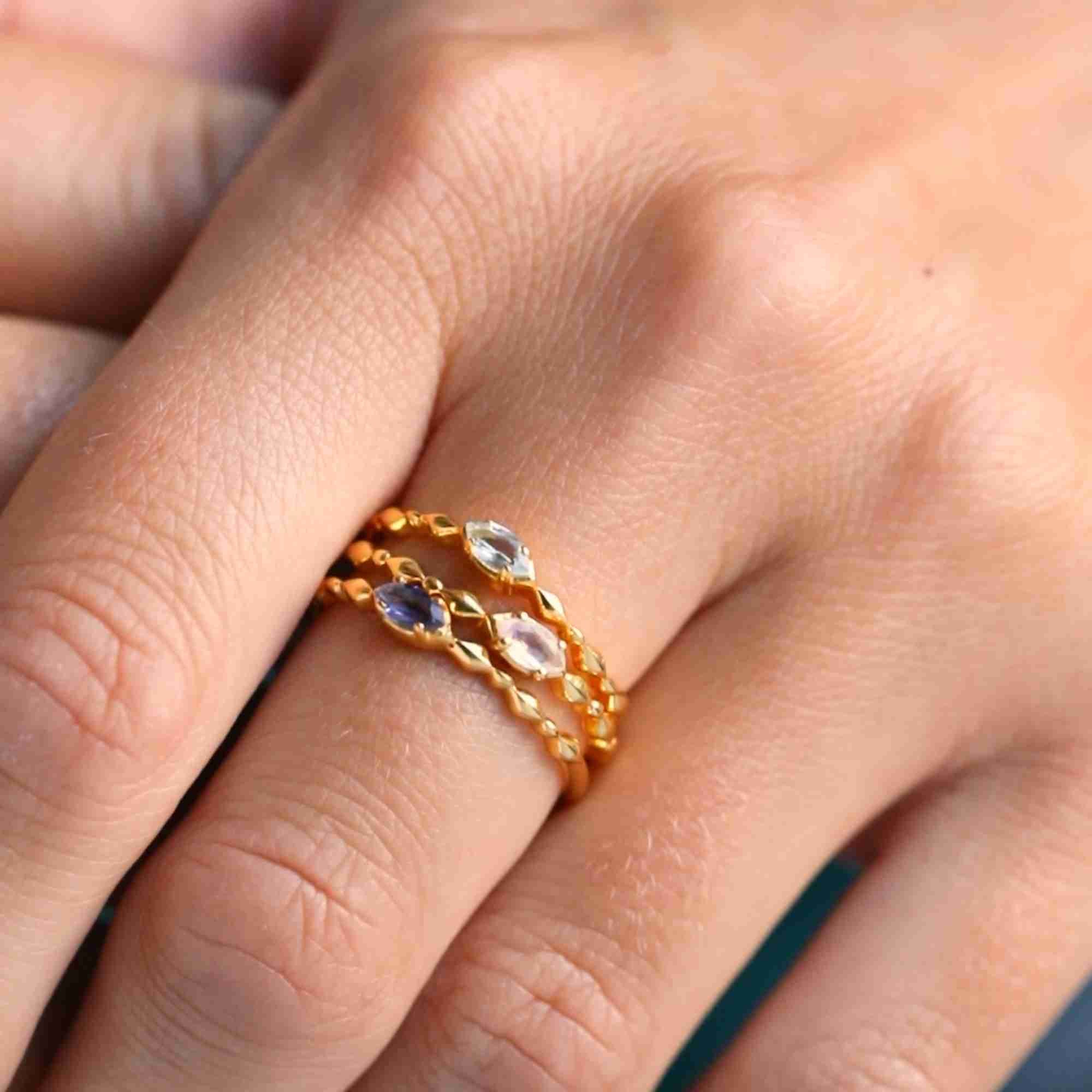 Close-up lifestyle portrait named Delicate Vintage showcasing three stacked dainty 22K gold vermeil rings on a model's finger. The minimalist stack highlights a slender ring featuring a horizontally set marquise-cut light blue Topaz gemstone on a textured, dotted bubble band by Protsaah Zurich.