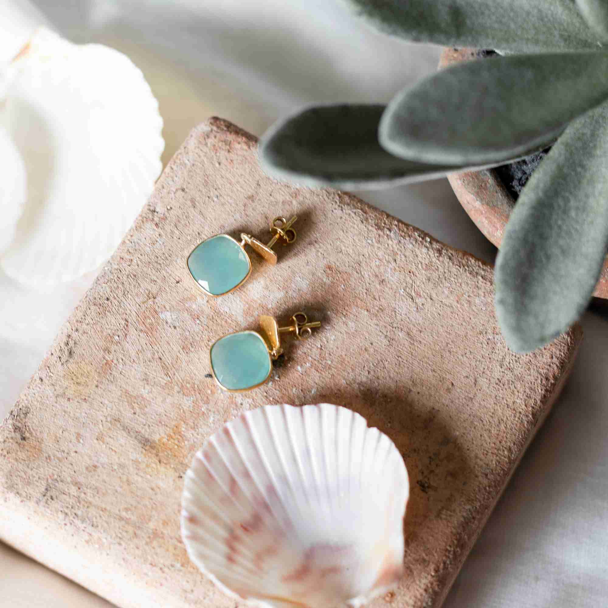 Lifestyle flatlay picture named Geo Glam displaying the Cushion-Cut Aqua Chalcedony Drop Earrings Gold. A pair of handcrafted 22K gold vermeil drop earrings featuring square, cushion-cut translucent sea-blue Aqua Chalcedony stones resting on a rustic terracotta block next to an open white seashell and green succulent leaves by Protsaah Zurich.