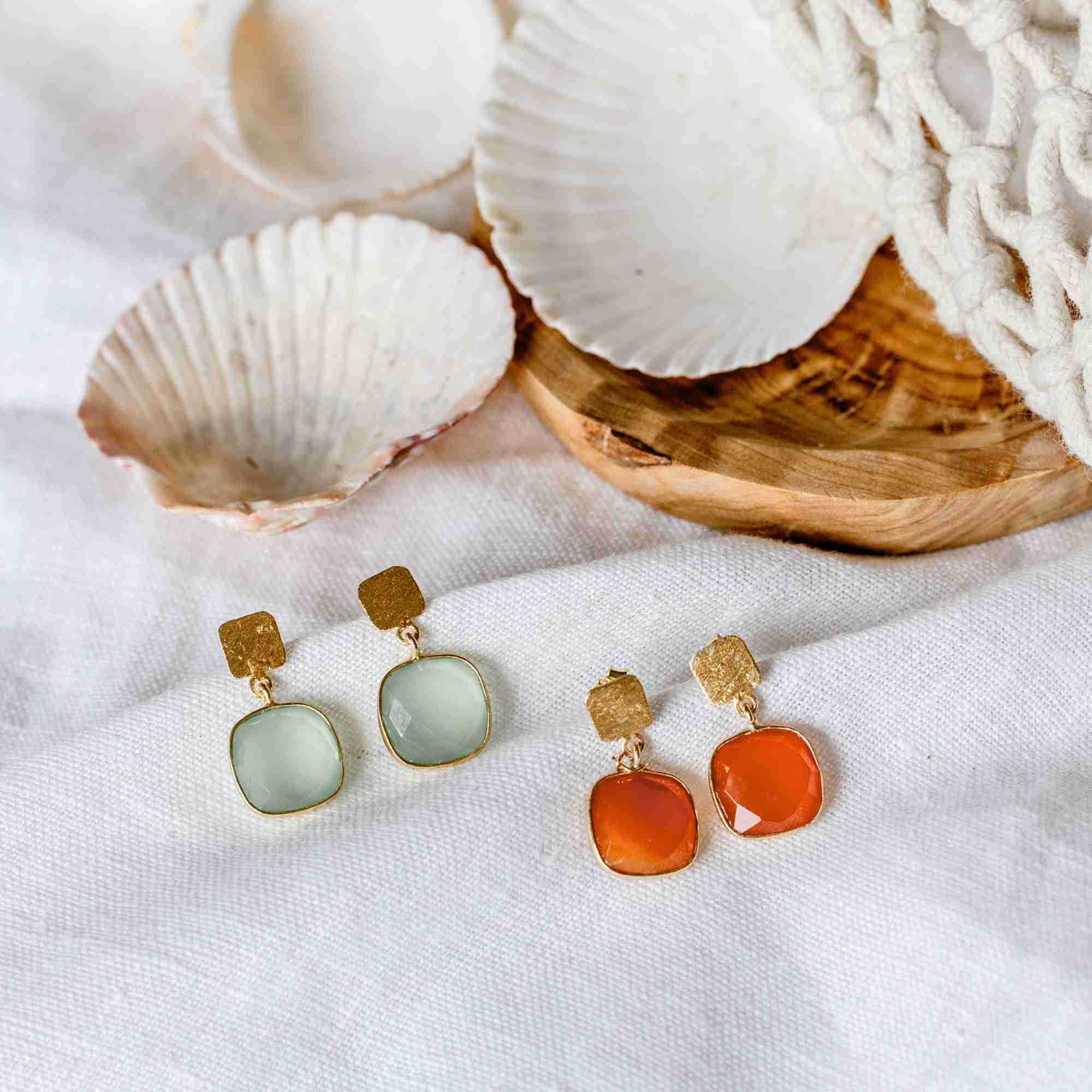Lifestyle flatlay picture named Geo Glam displaying the Cushion-Cut Carnelian Drop Earrings Gold alongside pale green prehnite drop earrings. The handcrafted 22K gold vermeil pairs feature square, cushion-cut translucent orange Carnelian gemstones suspended from textured gold studs, resting on textured white linen next to sea shells and a wooden plate by Protsaah Zurich.