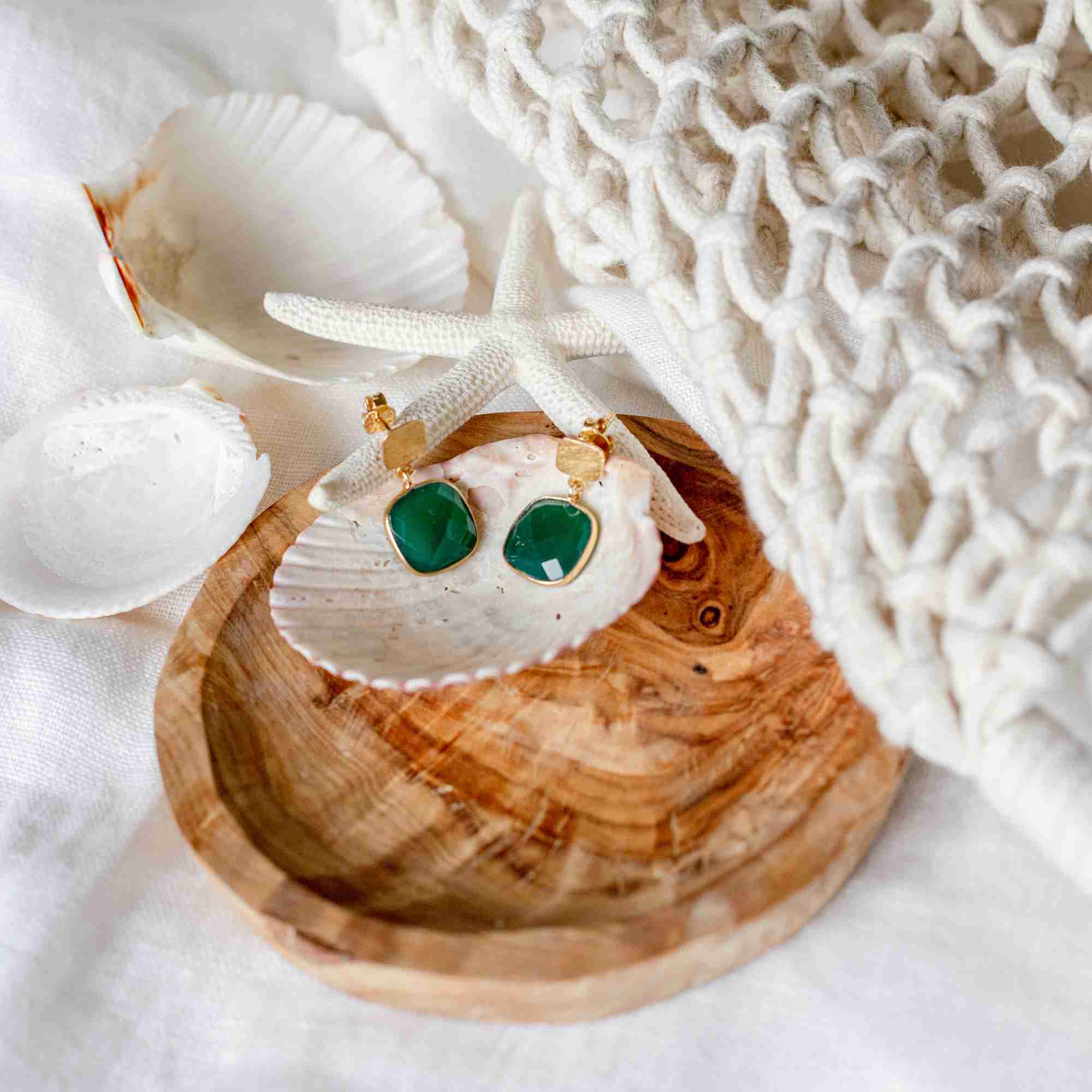 Lifestyle flatlay picture named Geo Glam displaying the Cushion-Cut Green Onyx Drop Earrings Gold. A pair of handcrafted 22K gold vermeil drop earrings featuring square, cushion-cut translucent emerald-green Onyx stones suspended from textured golden square studs, beautifully arranged inside a natural seashell on a carved wooden plate with a white starfish and a woven cotton net background by Protsaah Zurich.