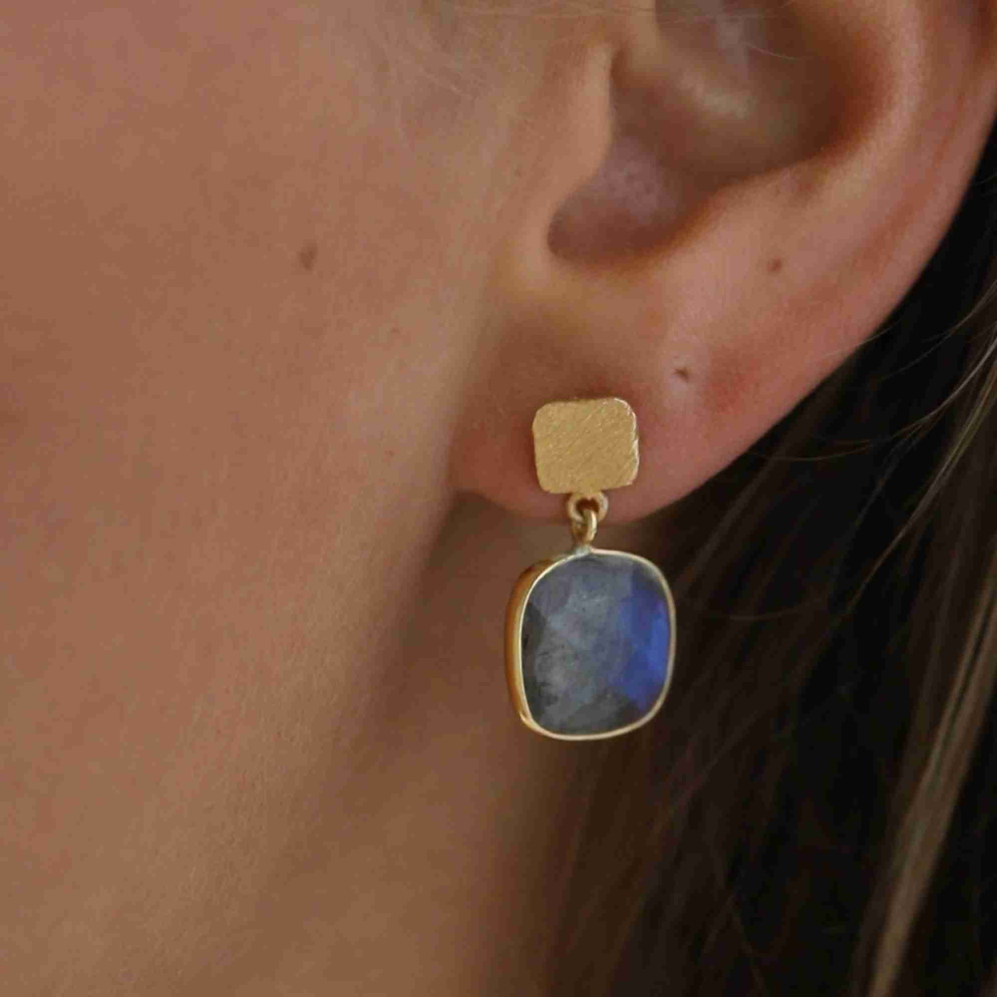 Detailed close-up lifestyle portrait named Geo Glam focusing on a model's ear wearing a handcrafted 22K gold vermeil drop earring, featuring a square, cushion-cut natural Labradorite gemstone with a brilliant iridescent blue flash suspended from a textured gold post by Protsaah Zurich.