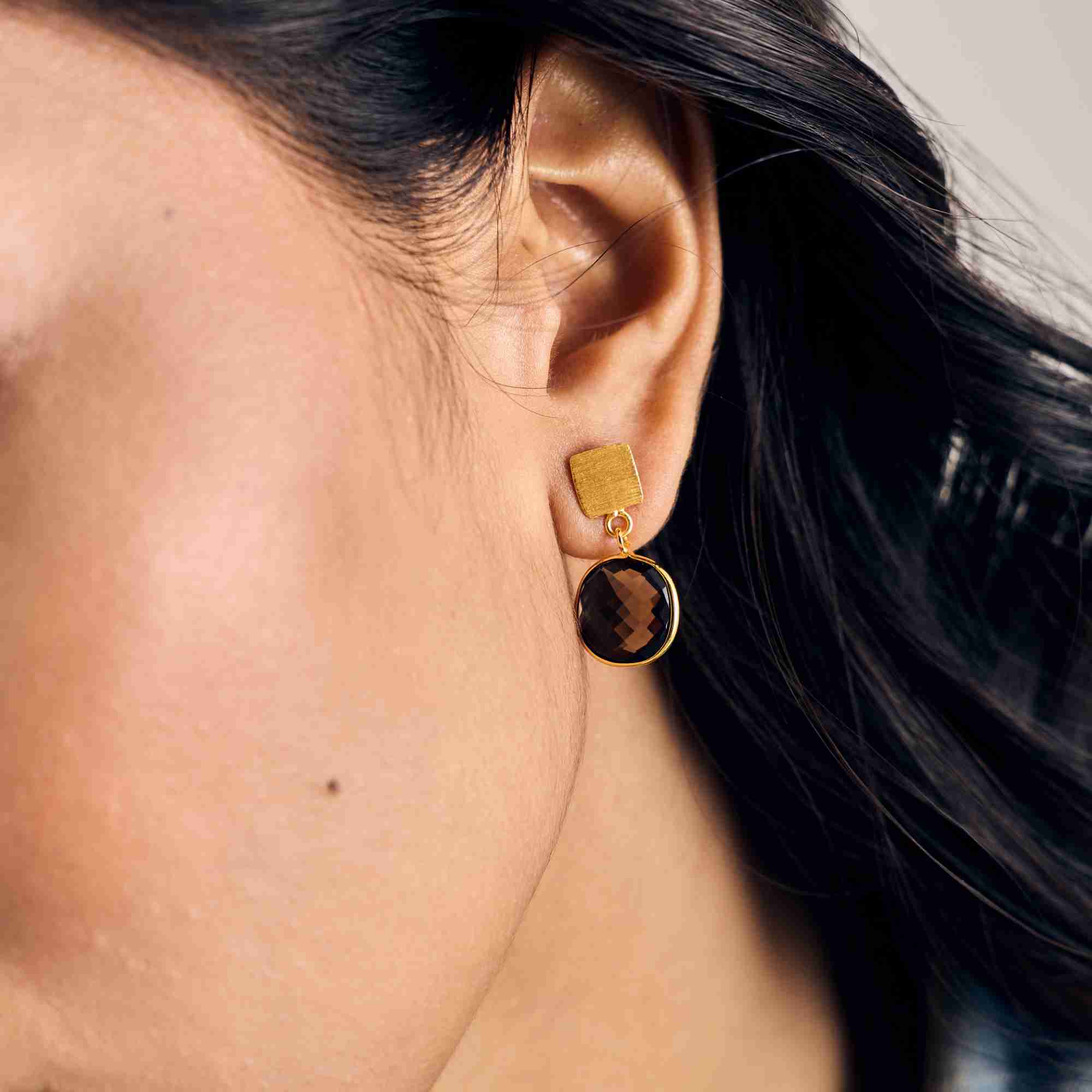Detailed close-up lifestyle picture named Geo Glam focusing on a model's ear wearing a handcrafted 22K gold vermeil drop earring set with a square, checkered cushion-cut translucent smoky-brown Quartz gemstone by Protsaah Zurich.
