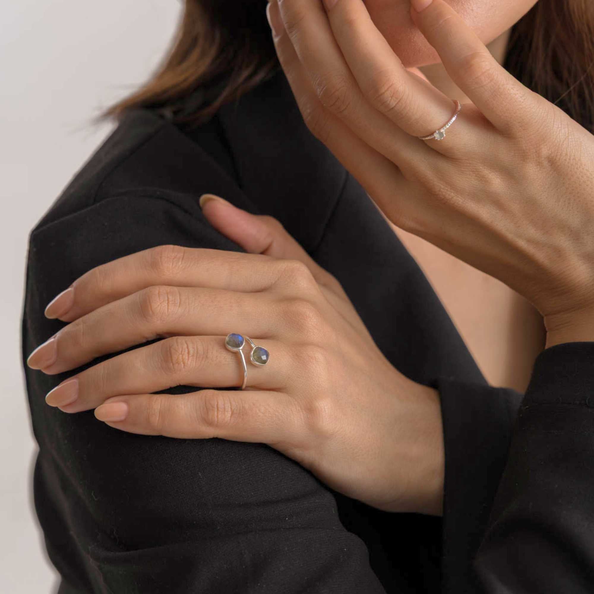 Lifestyle view of a model wearing a black blazer with her hand resting on her shoulder, showcasing the Geo Glam Toi et Moi Labradorite Ring Gold on her ring finger, referencing file name 50.jpg. The adjustable bypass band displays two iridescent, round bezel-set labradorite stones that capture soft flashes of blue.
