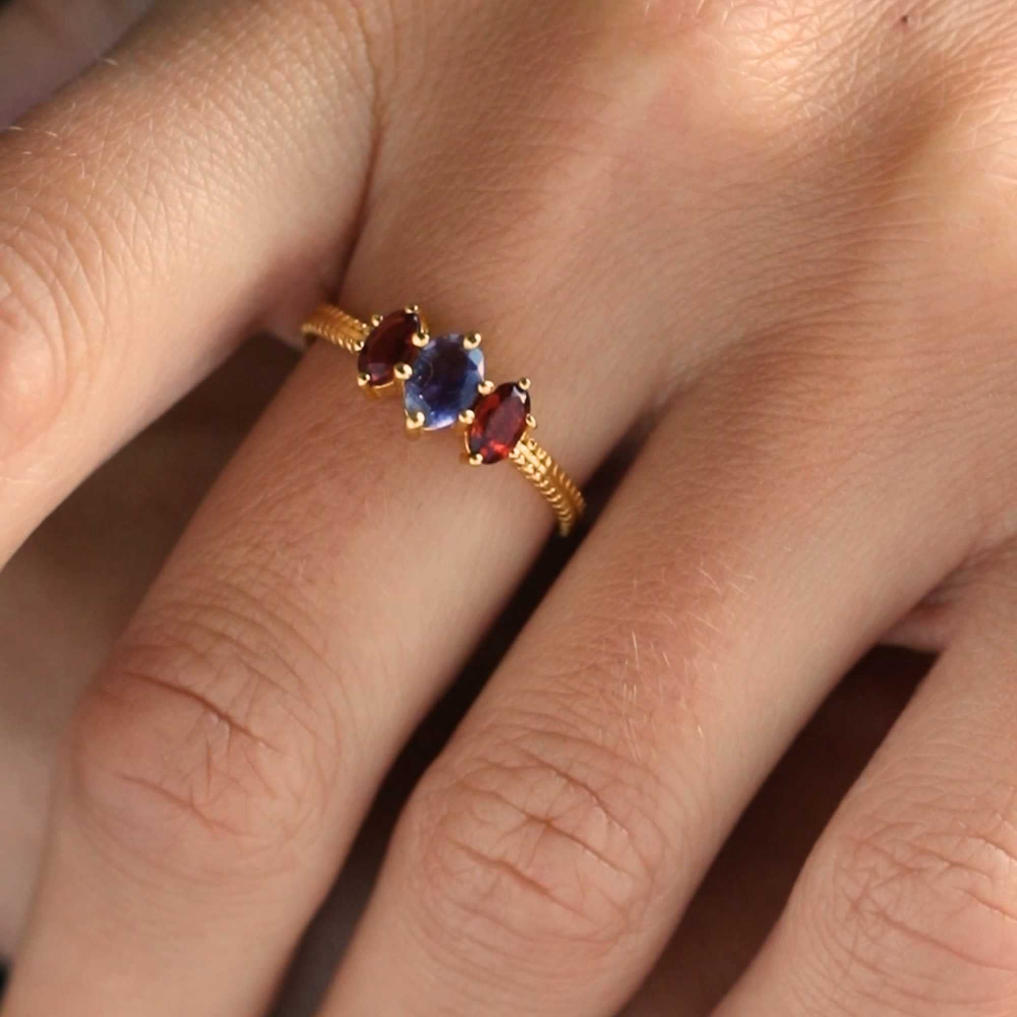 Macro close-up shot of the Kaleidoscope Iolite and Garnet Three-Stone Marquise Ring Gold being worn on a finger. The details highlight the prong-set violet-blue iolite stone framed by two rich red marquise garnets set against a textured gold band.