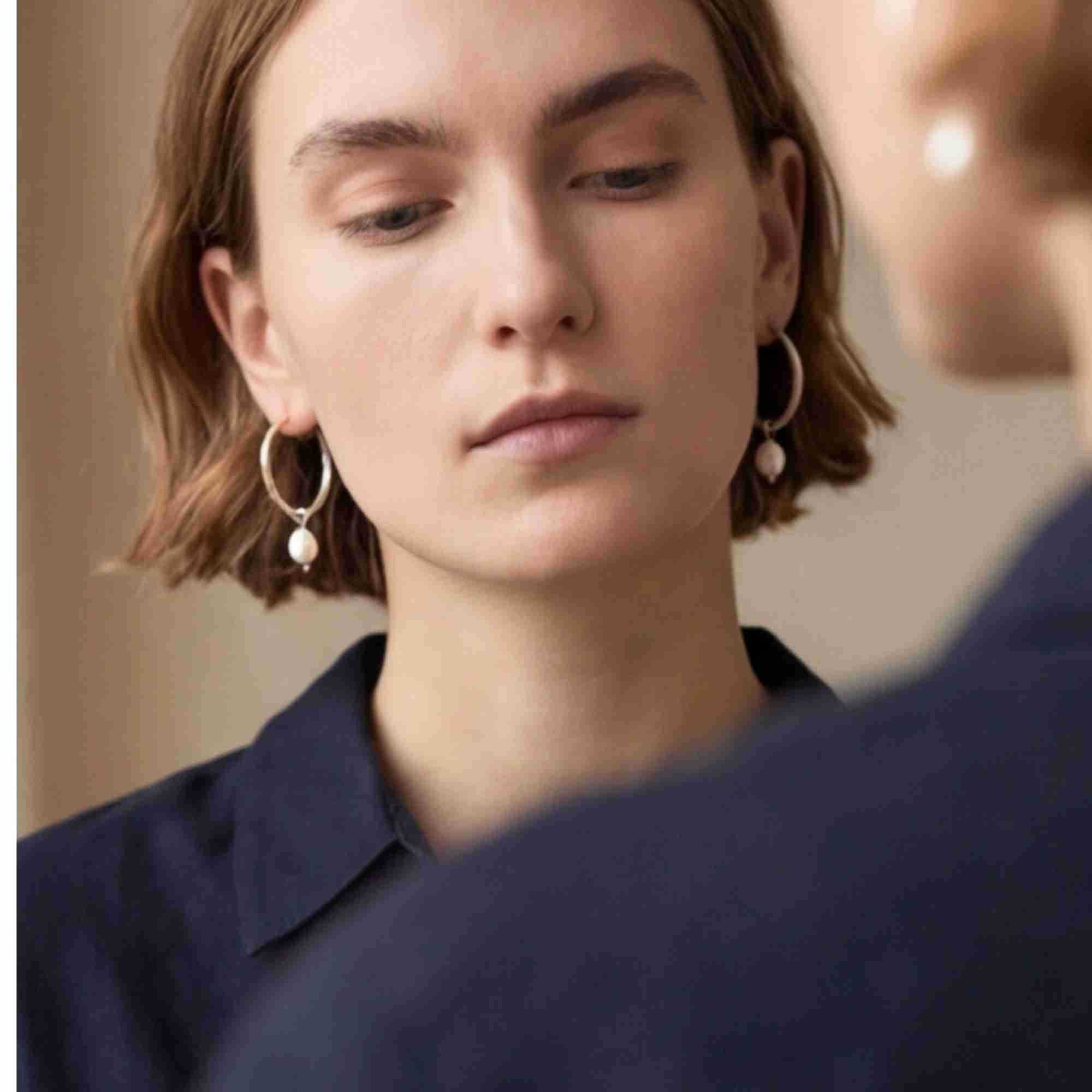 Lifestyle portrait of a model looking downwards while wearing the PEARLS GALORE Teardrop Hoop Earrings Silver, referencing file name Pearl_Galore_7.jpg. The shot, framed past another person in the foreground, highlights the elegant asymmetry and classic aesthetic of the textured silver hoops with hanging pearl charms on both earlobes.