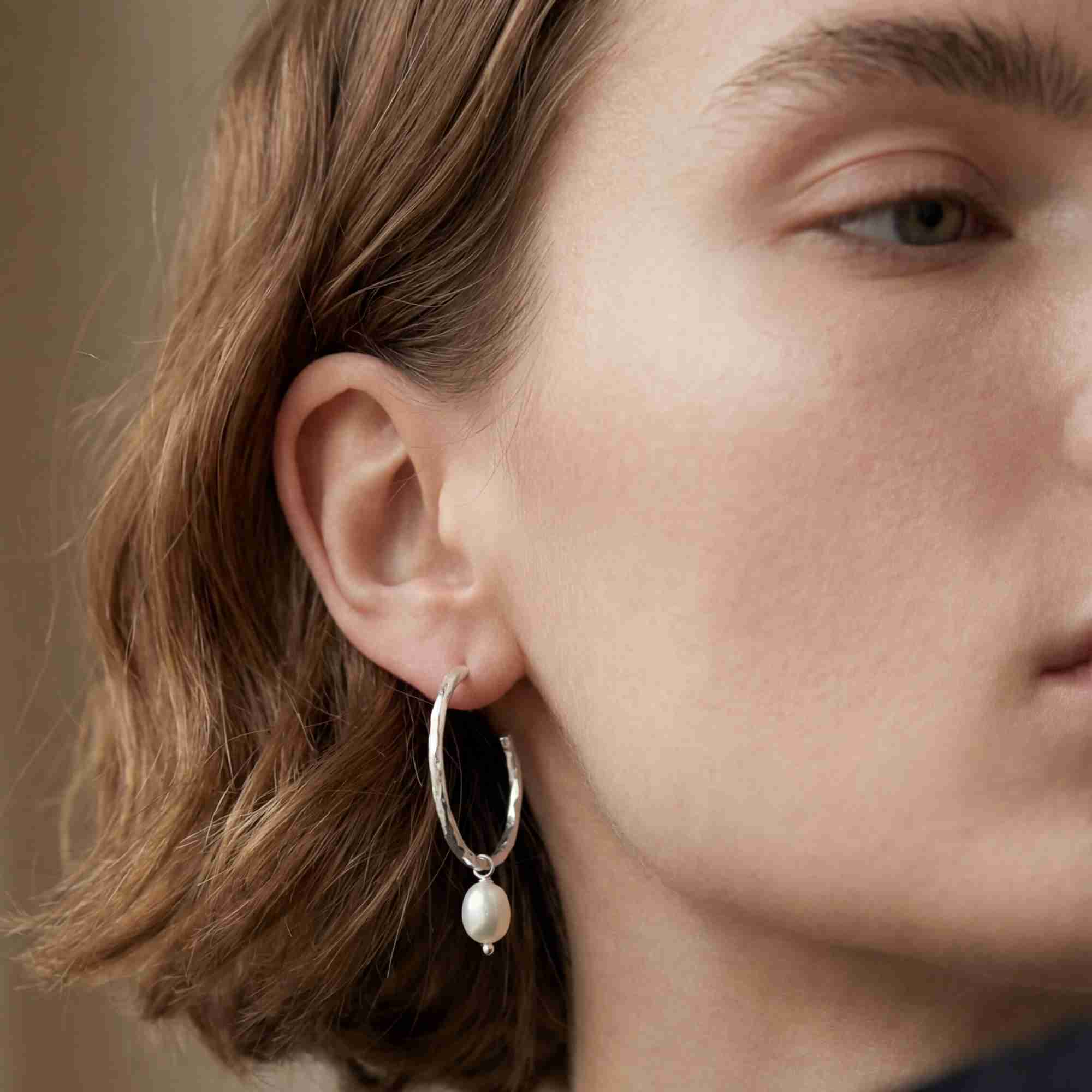Three-quarter lifestyle view of a short-haired model looking slightly away, showcasing the PEARLS GALORE Teardrop Hoop Earrings Silver, referencing file name Pearl_Galore_8.jpg. The neutral-toned lighting emphasizes the organic, hammered finish of the sterling silver hoop and the clean luster of the teardrop-shaped pearl.