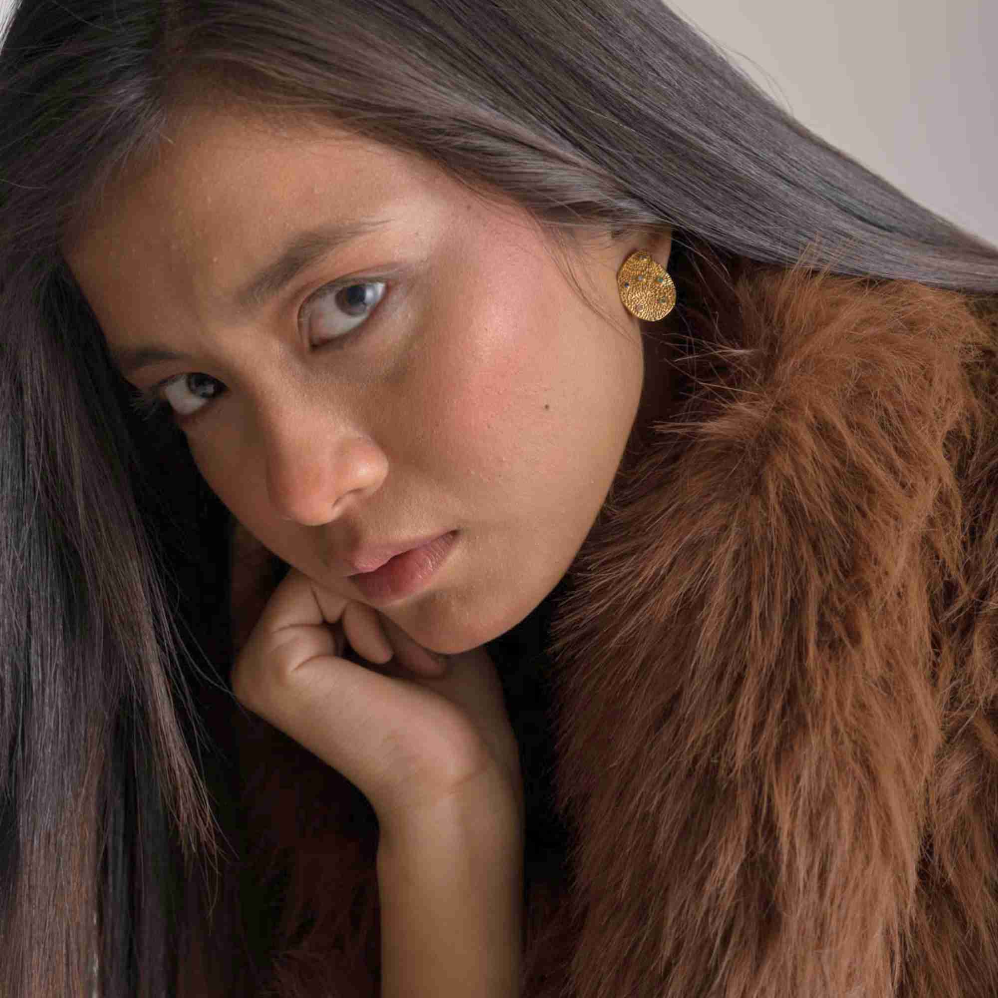 Candid-style lifestyle model image featuring the Ribbons and Lace Galaxy Multi-Gemstone Stud Earrings Gold, referencing file name Ribbon_Lace_18.jpg. The model tilts her head down slightly with a thoughtful expression, letting the textured golden stud earring catch the studio light above her faux-fur collar.