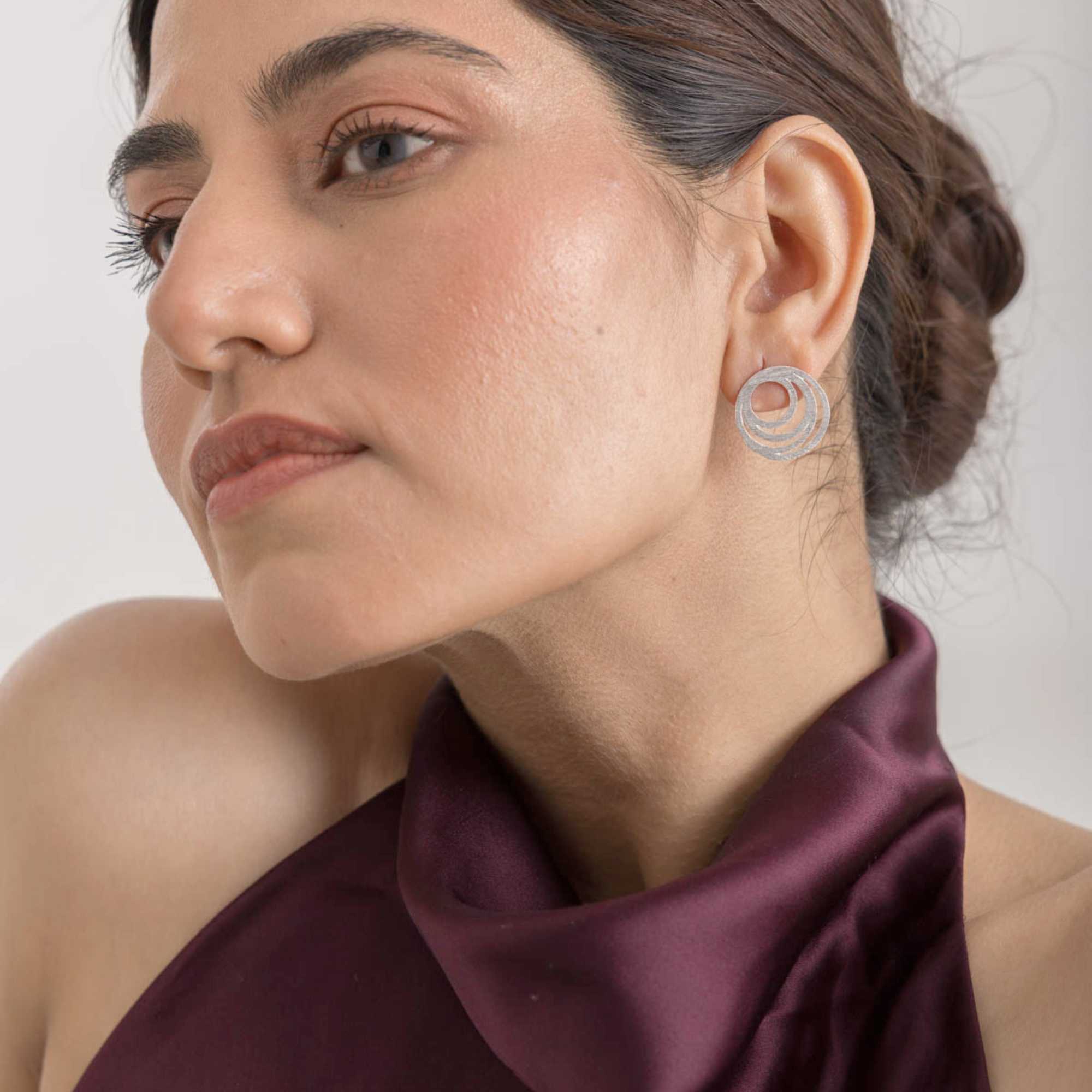 Lifestyle portrait shot of a model wearing the Shimmering Disks Intermingled Circles Stud Earrings Silver, referencing file name Shimmering Disks (70).jpg. Captured at an angled perspective, the image highlights the fine, shimmering scratch-finish texture across the interconnected silver circular bands, complementing her elegant updo hairstyle.