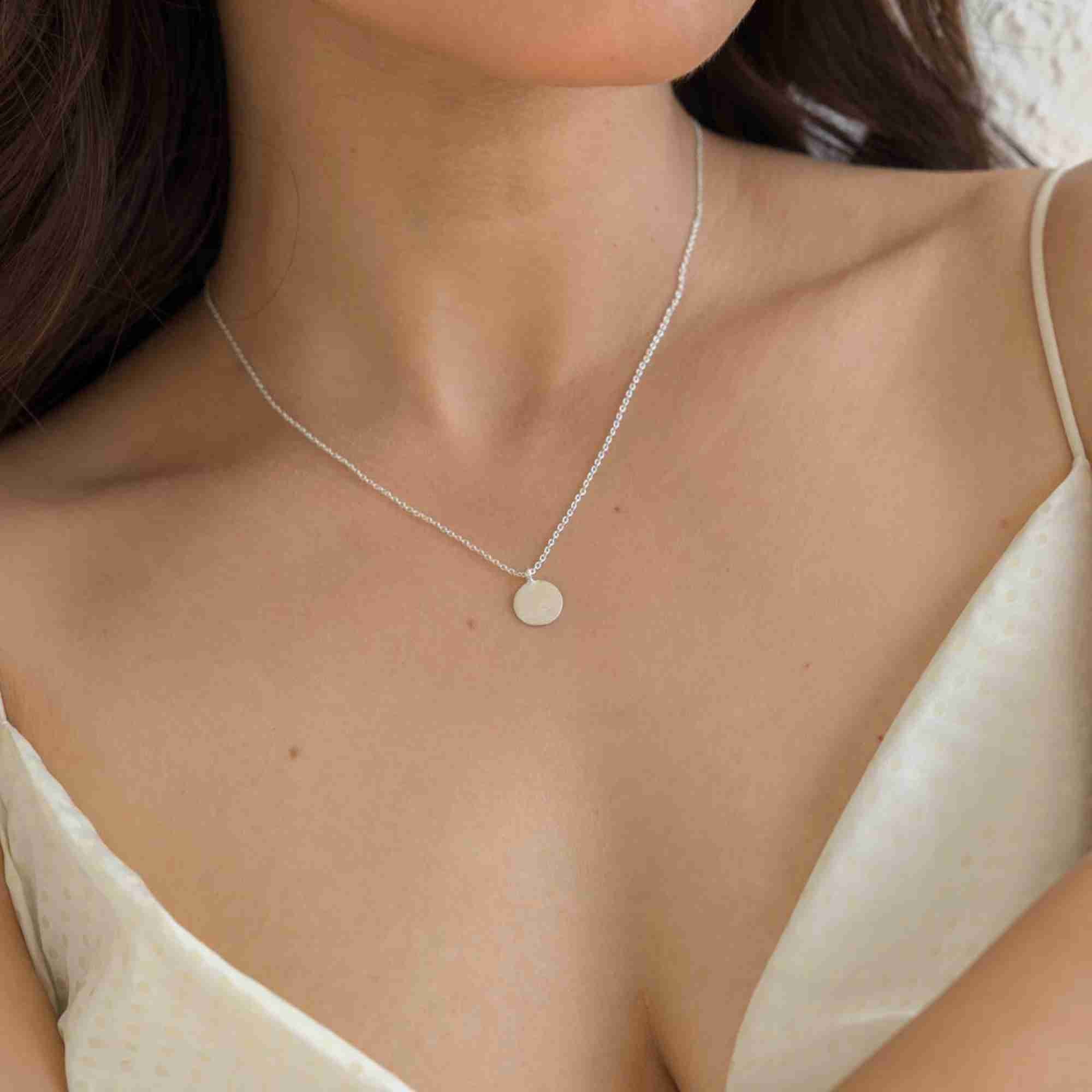 Lifestyle model view of the Shimmering Disks Minimalist Coin Pendant Necklace Silver, referencing file name 133.jpg. The shot shows a woman wearing a delicate, high-shine 92.5 sterling silver fine cable chain necklace featuring a flat, polished circular disk coin pendant that sits elegantly on her collarbone against a cream slip dress.