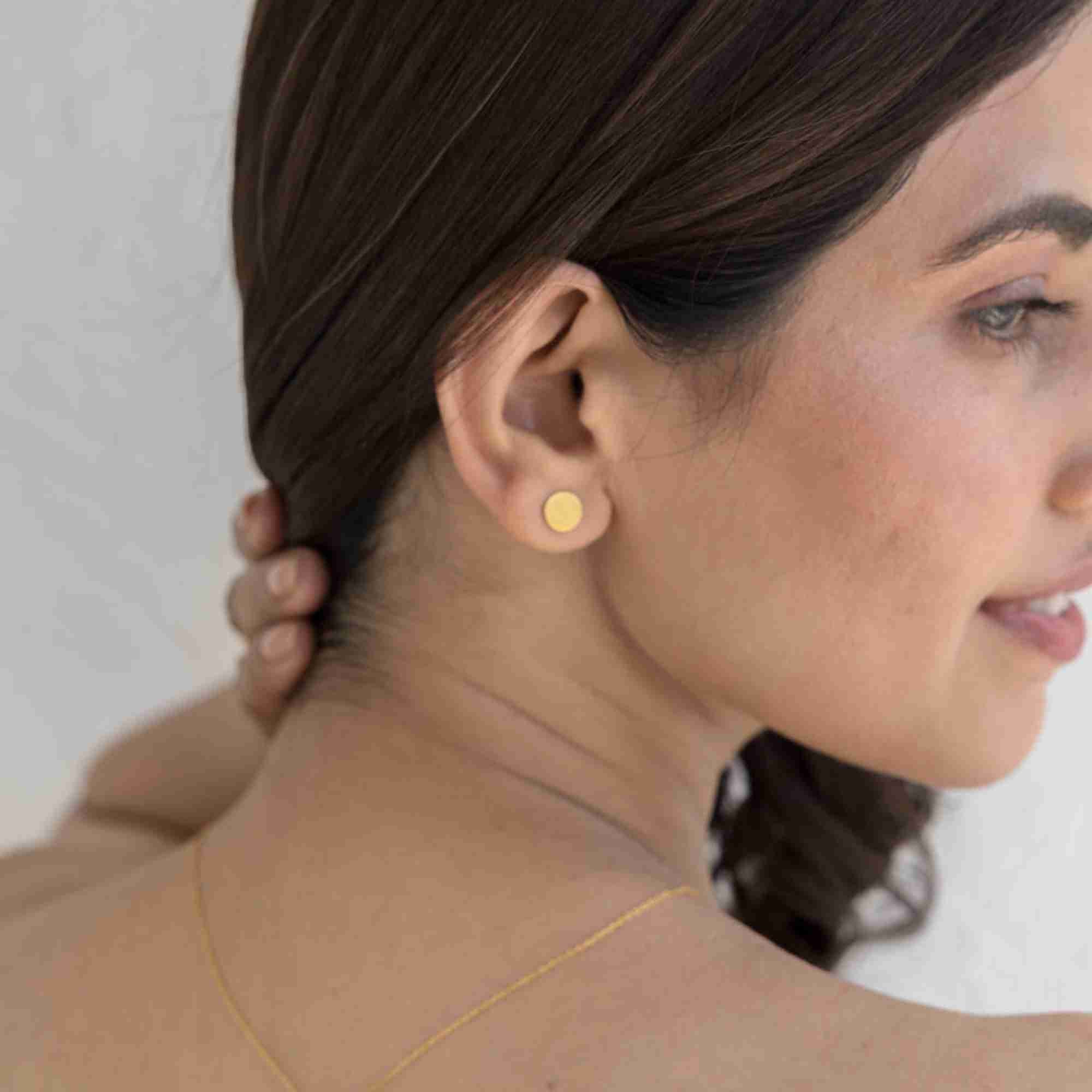 Angled rear lifestyle view of a woman modeling the Shimmering Disks Minimalist Stud Earrings Gold, referencing file name Shimmering_Disks_73.jpg. Posing with her hand gently touching the back of her neck, the image illustrates how the flat geometric gold disc stud sits elegantly against the ear.