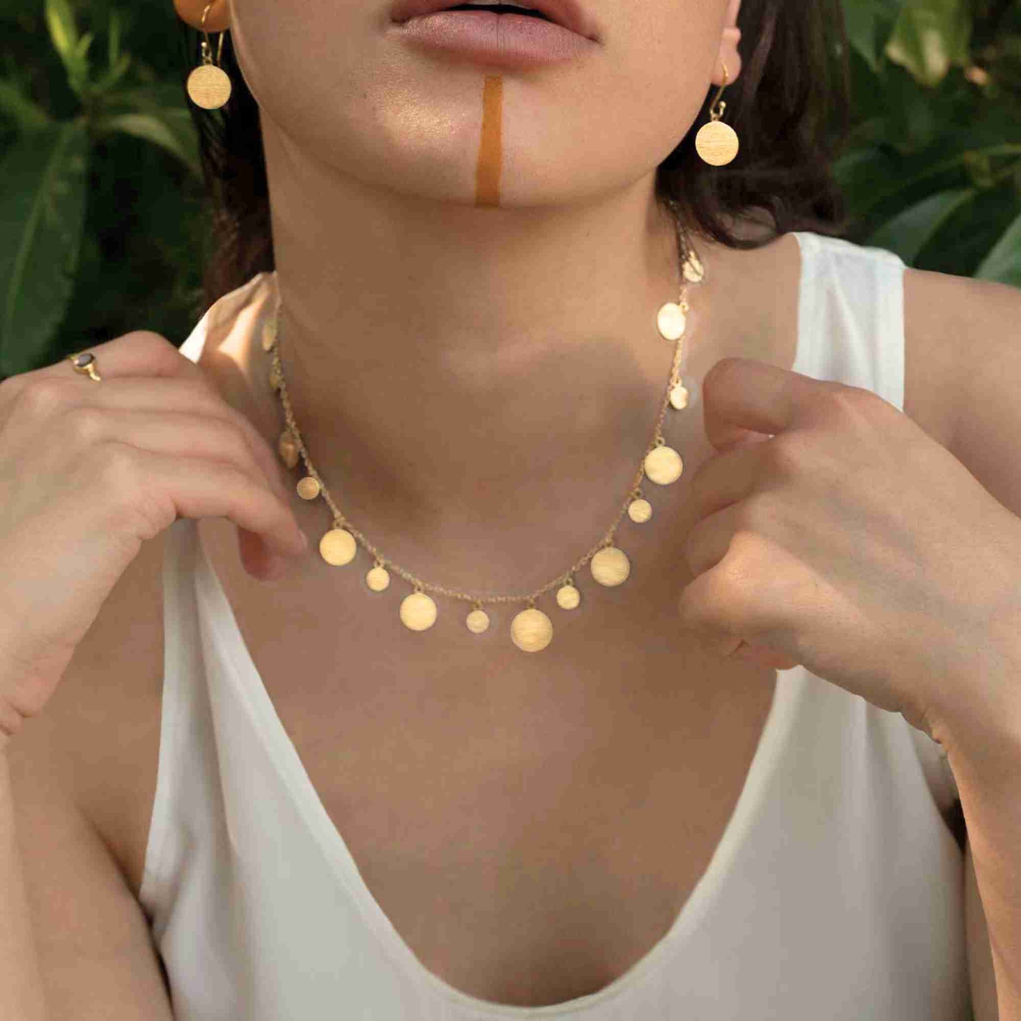 Frontal portrait view named Shimmering Disks (62).jpg showing a model adjusting the Shimmering Disks Scattered Statement Necklace Gold while wearing a white tank top and matching gold disc drop earrings against a lush green backdrop by Protsaah Zurich.